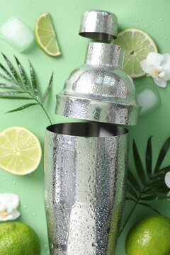 Shaker, limes, ice cubes, orchid flowers and leaves on green background, flat lay
