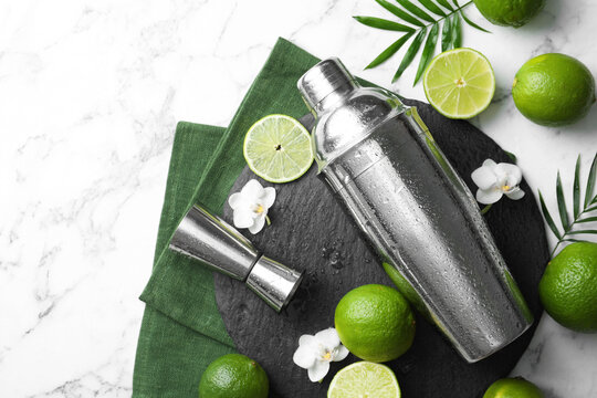 Shaker, limes, orchid flowers, leaves and jigger on white marble table, flat lay. Space for text