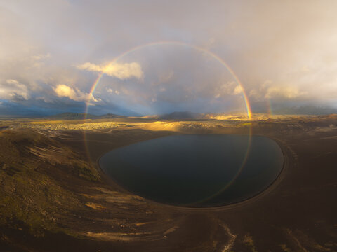 Aerial view of the Iceland Highlands featuring a volcanic crater lake illuminated by golden hour light with a full circular rainbow arching over the dramatic landscape in Iceland.