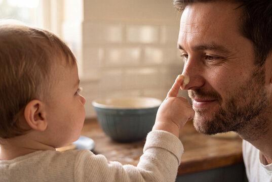 Cute baby playfully touching his smiling father's nose with cake batter while baking together in home kitchen