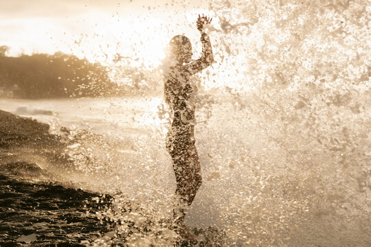 Silhouetted woman surrounded by ocean spray and golden sunset light on beach