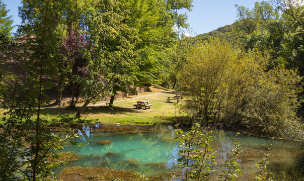 Aguas cristalinas y turquesas del nacimiento del r&iacute;o Ebro en Fontibre, rodeadas de frondosa vegetaci&oacute;n y un &aacute;rea recreativa con mesas de picnic.