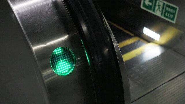Close‑up view of glowing green circular permission signal lamp on metal escalator side panel, showing traffic direction for orderly passenger movement in public transit space, exit safety sign in back