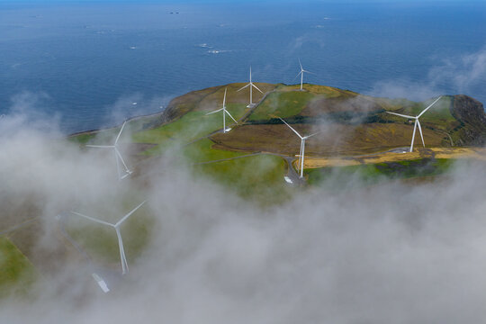 Aerial view of the Haramsoya wind farm with white turbines standing on a grassy coastal hill surrounded by fog and the sea in Haram, More og Romsdal, Norway.