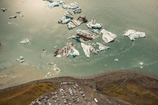 Aerial view of Jokulsarlon glacier lagoon with floating icebergs, dark volcanic sand beach, and a parking area with vehicles in Hofn, Sveitarfelagid Hornaf, Iceland.