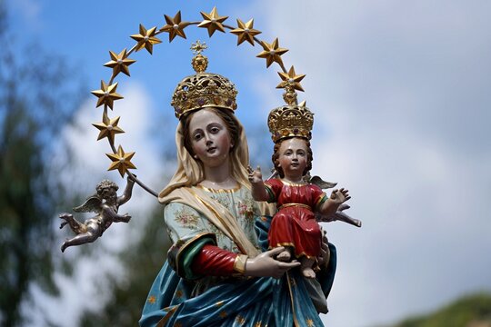 Statue of the Virgin Mary with the Infant Jesus. The Madonna and Child, crowned with a halo of stars and surrounded by cherubim, during a festive street procession under a blue sky.