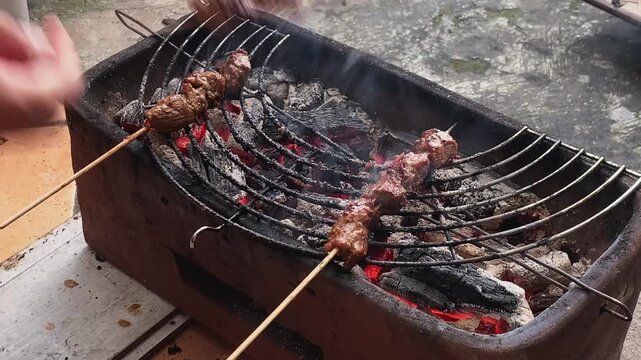 Hands turn skewers over glowing embers. Smoke rises from sizzling meat. Grill crackles with hot coals. Outdoor cooking with rustic charm. Perfect for food, festival, or backyard scenes.