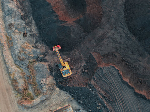 Aerial view of a yellow excavator working in a large coal mine with piles of black coal and tire tracks in the ground in Anggana, East Kalimantan, Indonesia.
