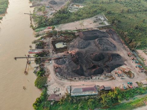Aerial view of a coal mine operation with large stockpiles of coal, conveyor belt systems, and heavy machinery situated along the riverbank in Anggana, East Kalimantan, Indonesia.