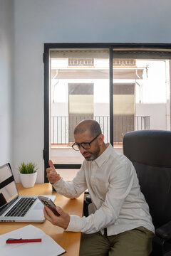 Confused man looking at smartphone beside laptop