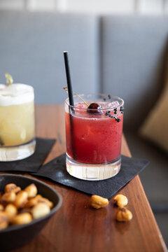 craft cocktail garnished with cherry on black napkin on wood table with fried corn kernels as snack in upscale bar