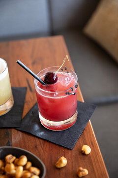 craft cocktail garnished with cherry on black napkin on wood table with fried corn kernels as snack in upscale bar