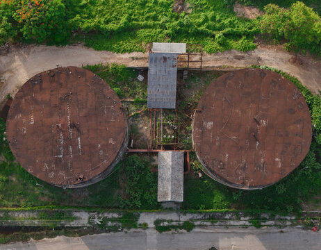 Aerial view of two large, weathered, circular industrial storage tanks connected by pipes, situated amidst greenery in San Pedro de Macoris, San Pedro de Macoris, Dominican Republic.