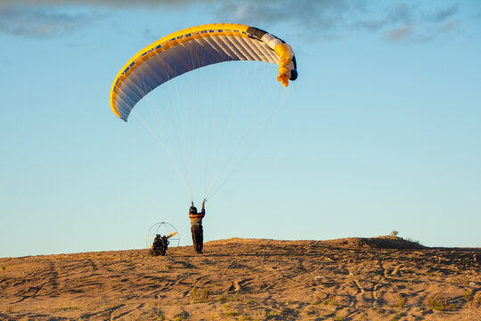 Paramotor pilot preparing yellow and grey paraglider wing on sandy ground against clear blue sky.