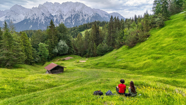 Two female hikers with backpacks walking on green alpine meadow towards mountain peaks, women friends trekking in the European Alps near traditional wooden hut, Bavarian Alps