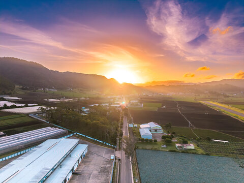 Aerial view of the sunrise over the agricultural landscape featuring greenhouses, cultivated fields, and rolling mountains under a vibrant sky in Constanza, La Vega Province, Dominican Republic.