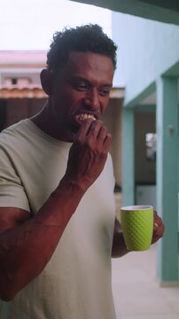 African American man standing in courtyard snacking and sipping from green mug with focused expression