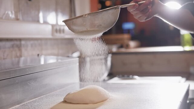 Slow motion baker sifting flour over dough for pizza on stainless countertop in bakery kitchen. handheld tracking pan.