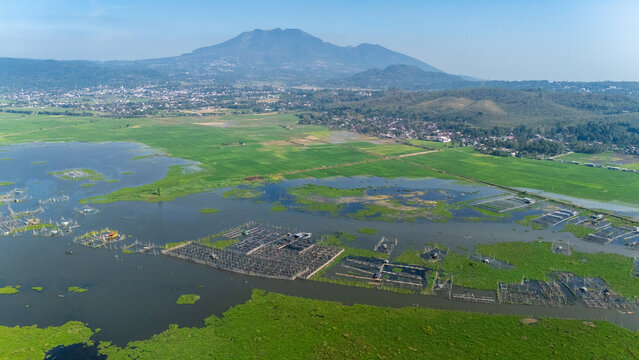Aerial view of fish farming cages in a lake surrounded by lush green rice fields and a distant town with a mountain in the background in Salatiga, Central Java, Indonesia.