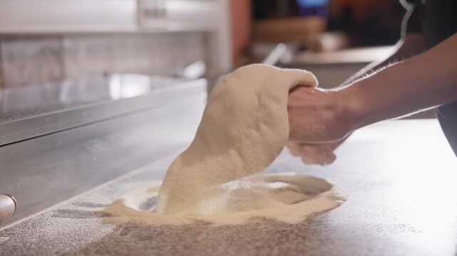 Handheld close up of baker shaping pizza dough on floured countertop in restaurant kitchen.