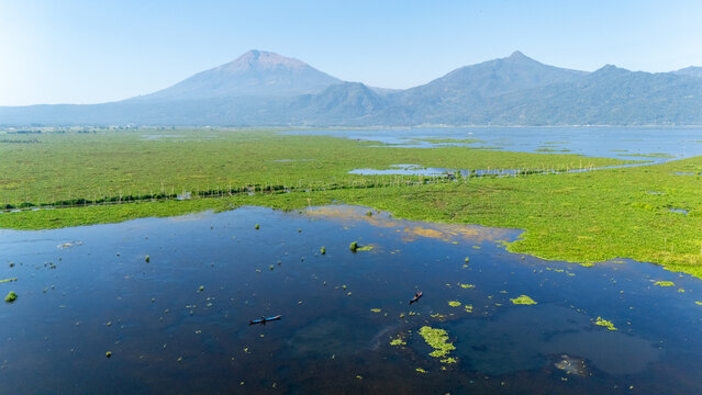 Aerial view of Rawa Pening lake with lush green water hyacinths, small fishing boats on the water, and mountains in the background in Salatiga, Central Java, Indonesia.