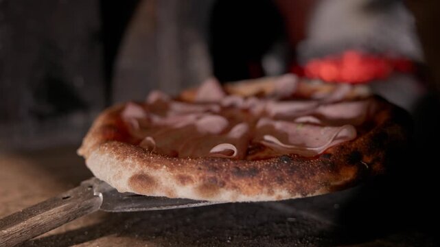 Panning shot pizza being slid into wood fired oven on peel close up showing crust charring and melting cheese.