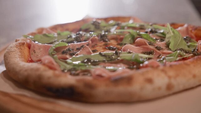 Slow motion closeup of pizza garnished with arugula and ham being placed on wooden table in restaurant tabletop tracking shot.