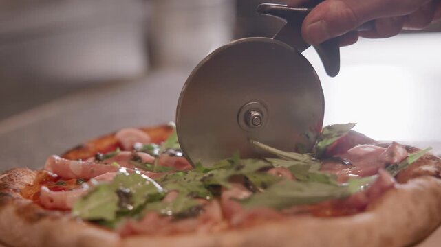 Closeup of hand slicing pizza with wheel cutter on wooden board at kitchen countertop near oven and prep area.