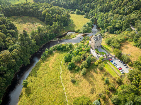 Aerial view of Neidpath Castle situated on the banks of the River Tweed, surrounded by lush green fields, dense woodland, and a small parking area in Peebles, Scotland, United Kingdom.