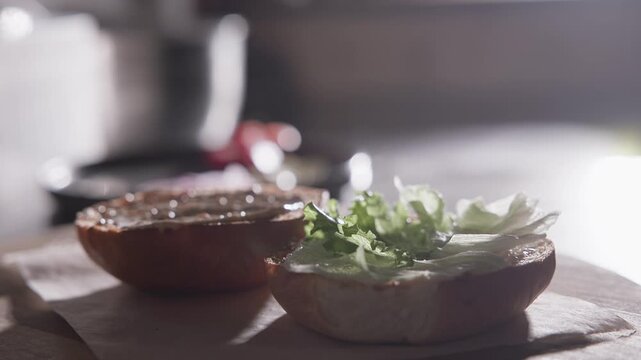 Slow motion hand placing lettuce and onion on burger bun in kitchen prep area with backlit soft light.