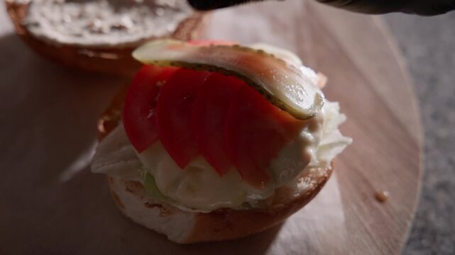 Slow motion hand placing pickle slices on open sandwich with tomato and lettuce on wooden board in kitchen.
