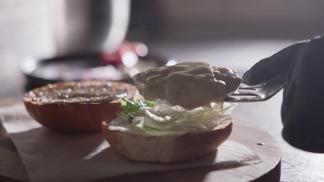 Tracking shot of cheeseburger patty being placed and cheese melting on open bun over wooden countertop in kitchen.