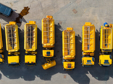 Aerial view of a fleet of yellow maintenance trucks parked in a row on a paved surface, with one truck equipped with a snowplow blade in the United Kingdom.