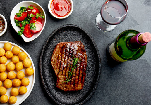 Grilled beef steak served on cast iron plate with red wine, tomato salad and potatoes ball.  Barbecue, bbq meat beef tenderloin. Top view, slate background