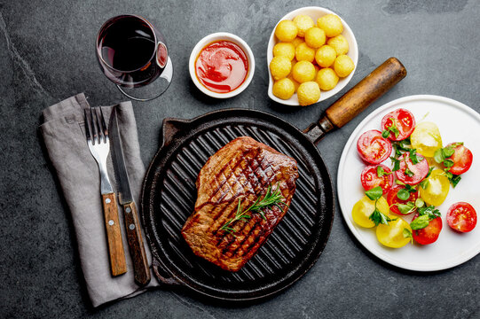Grilled beef steak on grill pan served with tomato salad, potatoes balls and wine. Barbecue, bbq meat beef tenderloin. Top view, slate background