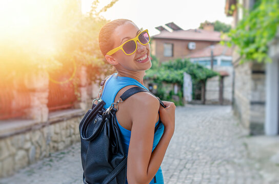 A cheerful young woman wearing yellow sunglasses and a backpack smiles while exploring an old sunny street on a bright day.