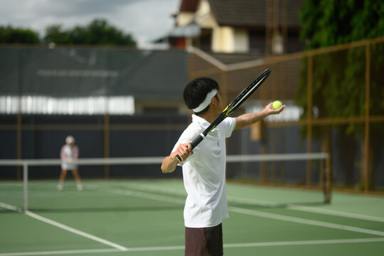 Rear view of Young Male Tennis Player Preparing to Serve on Outdoor Green Court with Opponent in Background