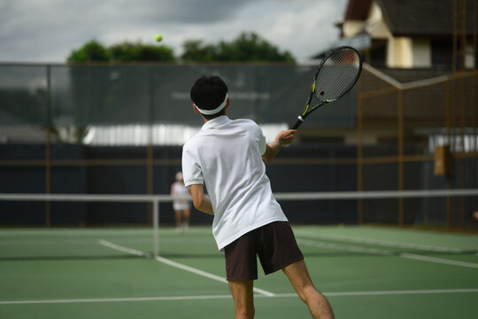 Rear view of Young Male Tennis Player Hitting Ball on Outdoor Green Court During Cloudy Day