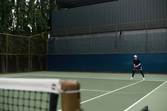 Young Asian Tennis Player in Ready Position on Outdoor Hard Court with Net in Foreground