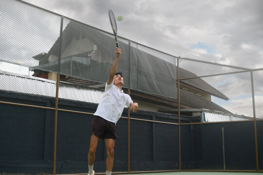 Young Asian Man Serving Tennis Ball on Outdoor Court with Cloudy Sky Background