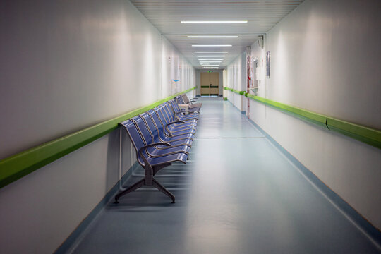 Empty illuminated hospital corridor featuring a long row of empty blue waiting chairs along the wall leading towards closed double doors at the end of the medical facility hallway.