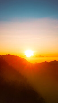 Aerial view of Mount Bromo at sunrise with golden light illuminating the volcanic landscape and mountain silhouettes in Tosari, East Java, Indonesia.