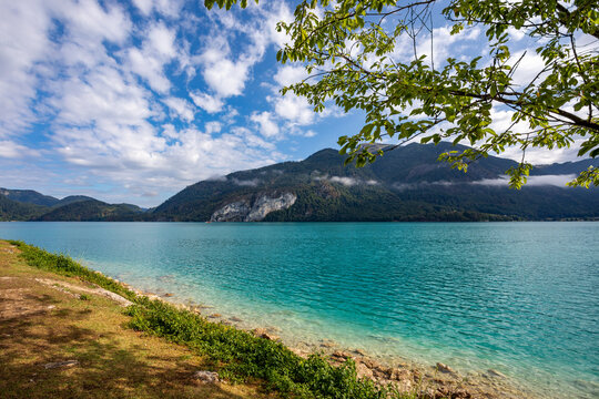 Turquoise Wolfgangsee lake with distant mountains under a cloudy sky and foreground foliage in Austria