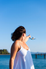 A woman points toward an approaching airplane near the coast in Kerkira, Greece. She stands outdoors near the blue sea under a clear sky. © dsbedarieva