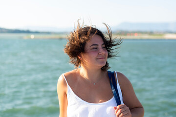 A young woman stands outdoors near the sea in Kerkira, Greece. The wind blows her short dark hair as she looks off to the side on a sunny day. © dsbedarieva