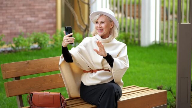 Smiling woman on wooden bench films herself with smartphone and greets someone off screen. She keeps cheerful, welcoming expression while sitting outside with her bag nearby, waving with raised hand.