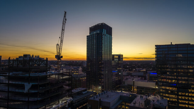 Aerial view of Kendall Square featuring modern glass skyscrapers, a construction site with a tall crane, and a vibrant sunset sky in Cambridge, Massachusetts, United States.
