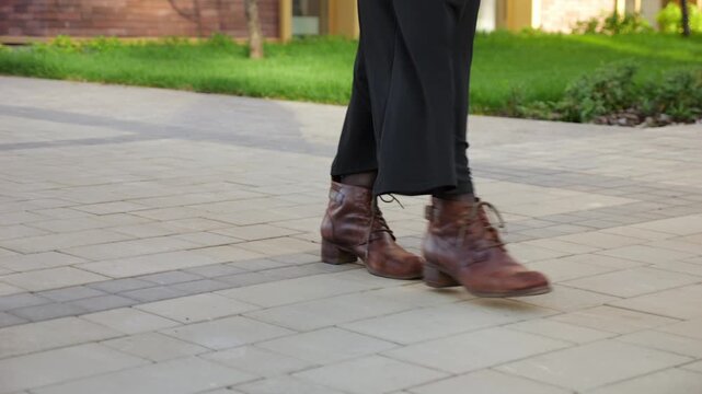Person in black skirt steps forward in brown lace up boots on stone path. Walking motion feels calm and purposeful, with close view of footwear against clean courtyard paving and lawn edge.