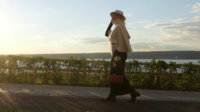 Woman in stylish outfit walks along waterfront path, carrying handbag and touching her hat. Fashion stroll at golden hour pairs calm movement with warm light and composed, elegant mood.