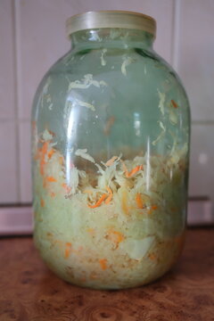 Homemade sauerkraut with shredded carrots sits inside a clear glass jar, preparing for fermentation on a wooden kitchen counter during the day.